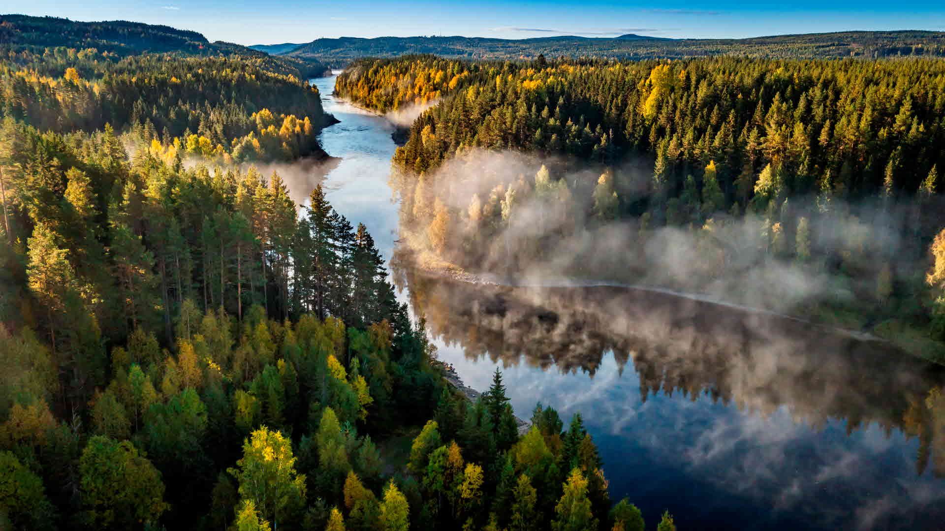 Flygbild tagen över en svensk skog, med en å som löper från det nedre högra hörnet av bilden upp längs med skogen. Låga moln hovrar över sjön.