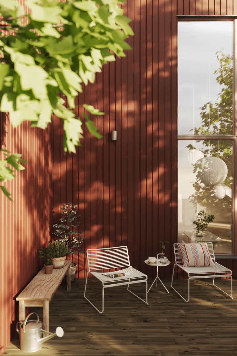 Närbild på ett hus, klädd i en rödmålad ytterpanel. I förgrunden två utomhusfåtöljer och ett sidobord.