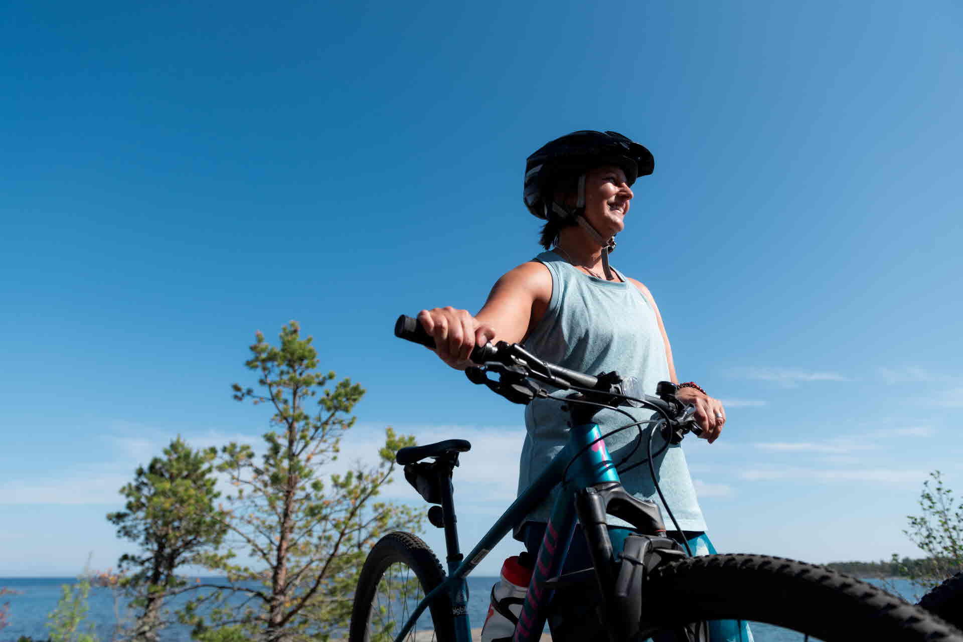 A person standing next to a bicycle, wearing a bike helmet, with the ocean in the background.