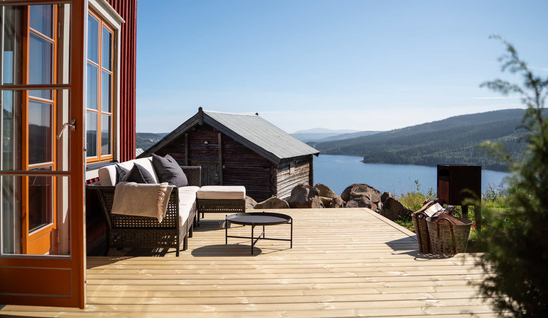 En trall intill ett rött hus med senapsgula fönsterfoder. På trallen står en soffgrupp och i bakgrund ser man en stor sjö och berg.