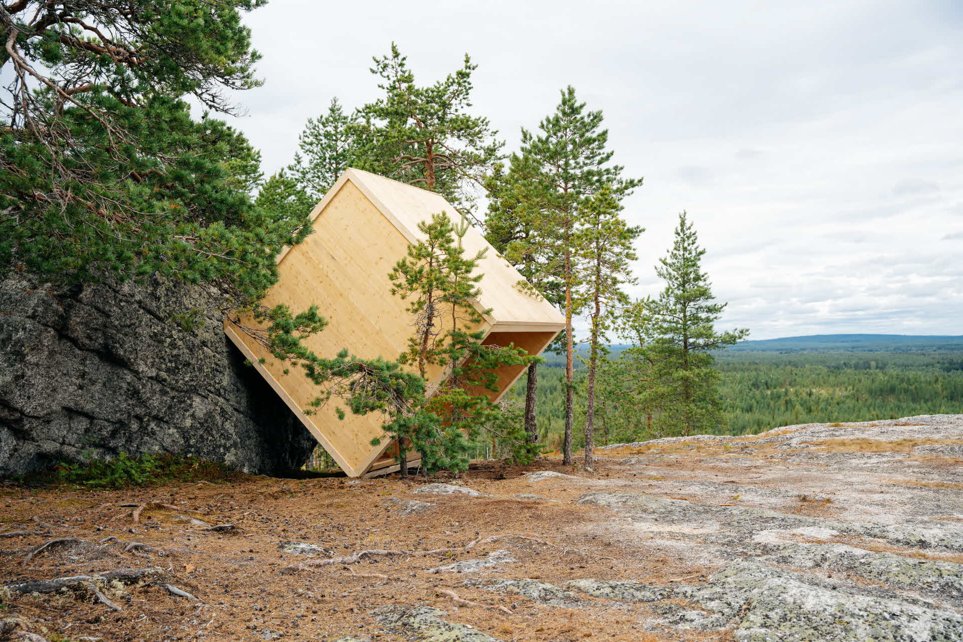 Ett vindskydd i trä format som en kub, står lutat i 45 grader mot ett stenblock.