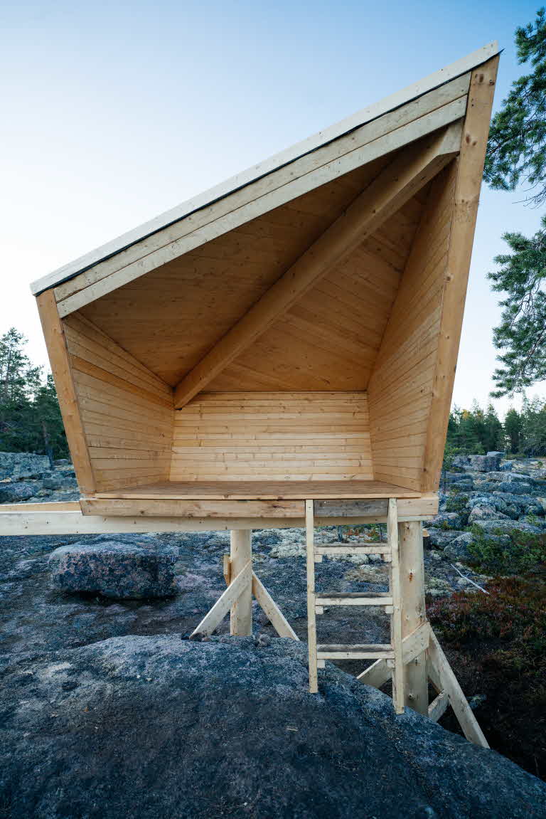 En gapahuk i tre, plassert blant store steinblokker og furutrær.