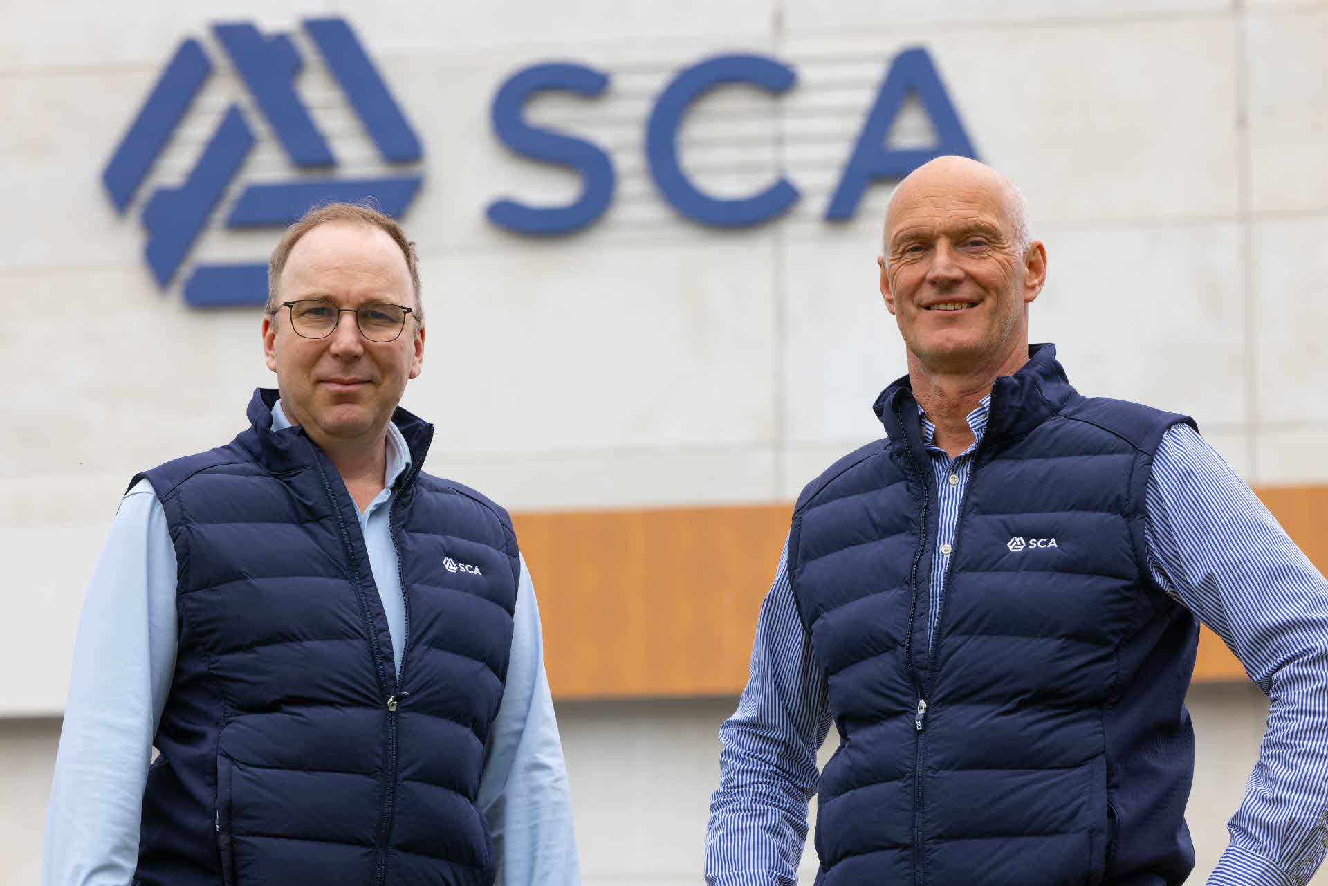 Two men standing in front of a building with SCA logo on it