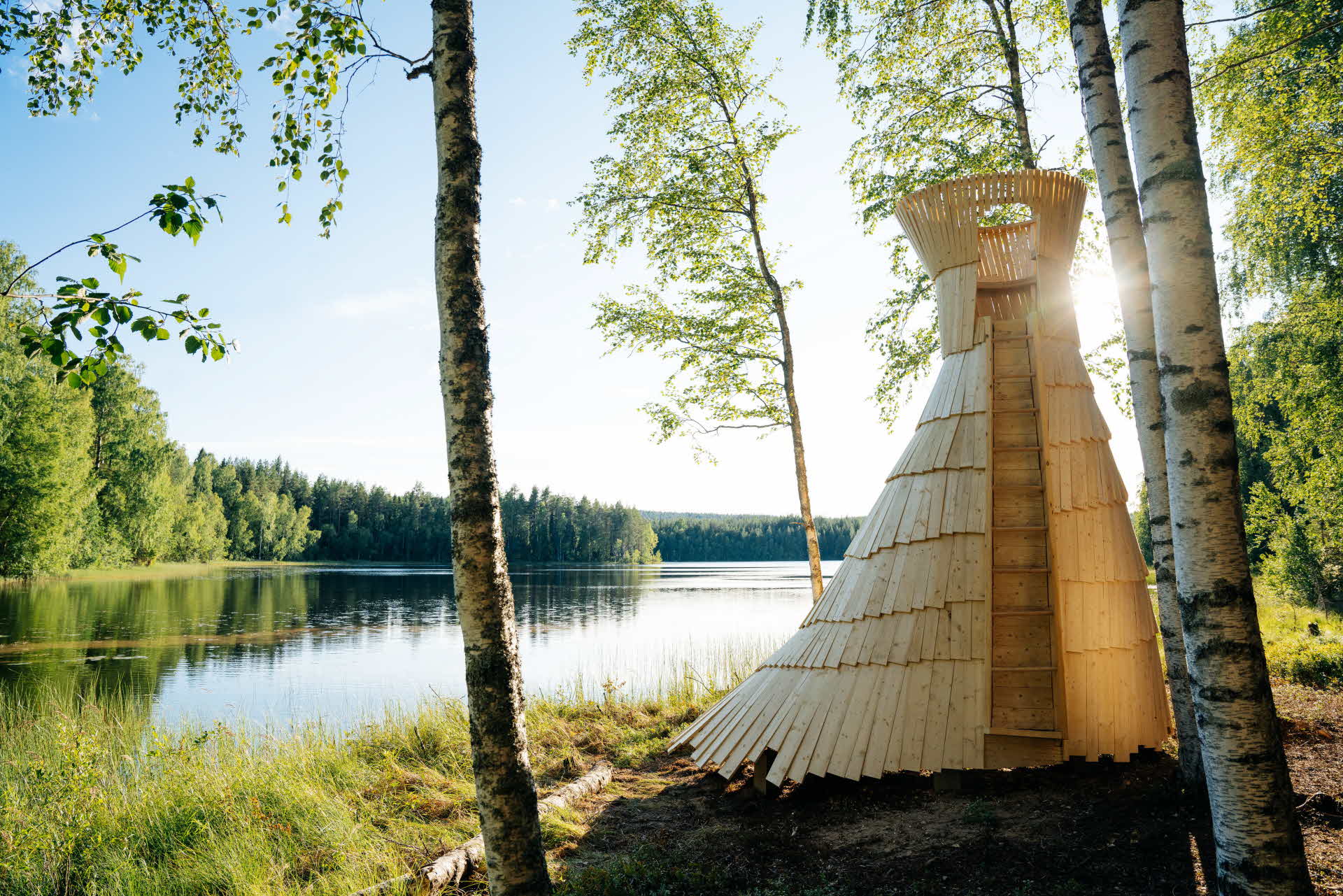 Ett högt vindskydd placerat framför en sjö, omringat av björkar.