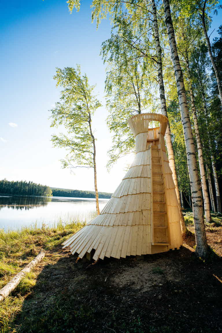 Ett högt vindskydd i trä. I bakgrunden grönskande björkar och en sjö.