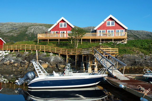 Vy från havet inöver brygga med båt och två röda stugor.