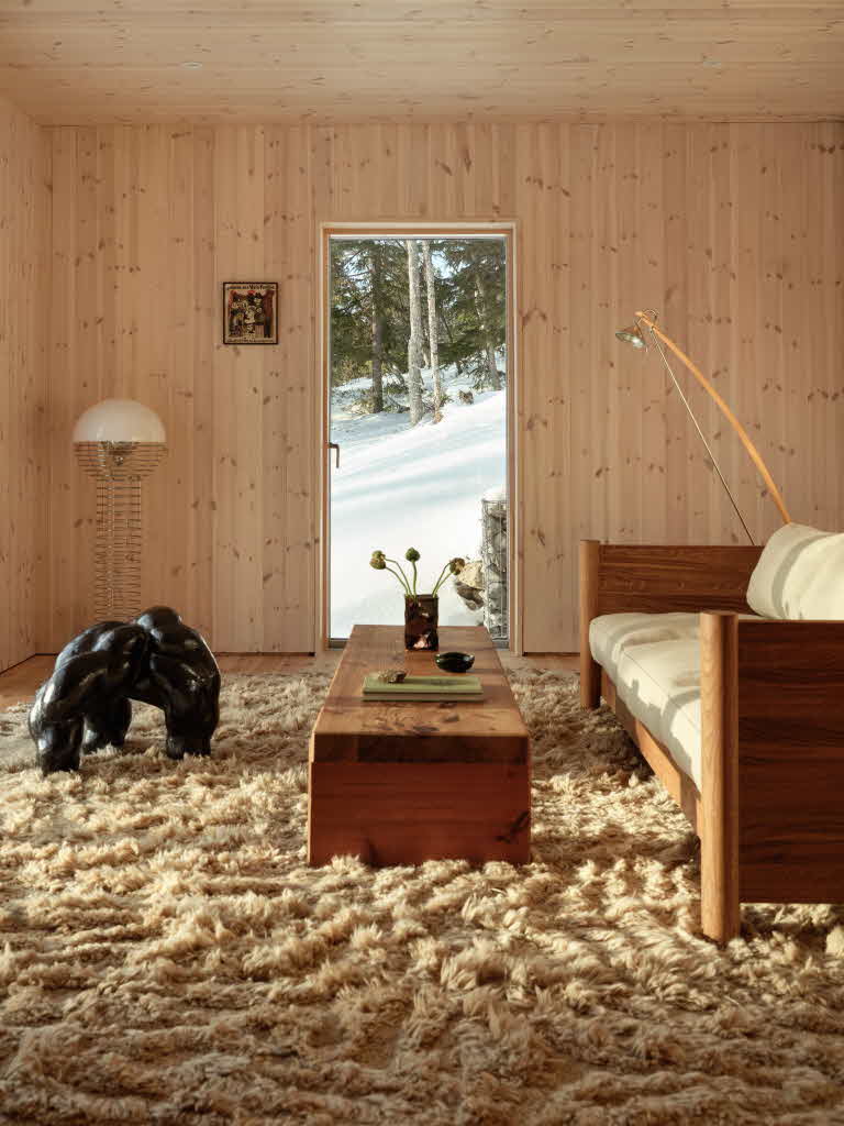 Vardagsrum med en fluffig matta på golder, ett lågt bord och en soffa i trä, ett stort fönster där man ser snön utanför.