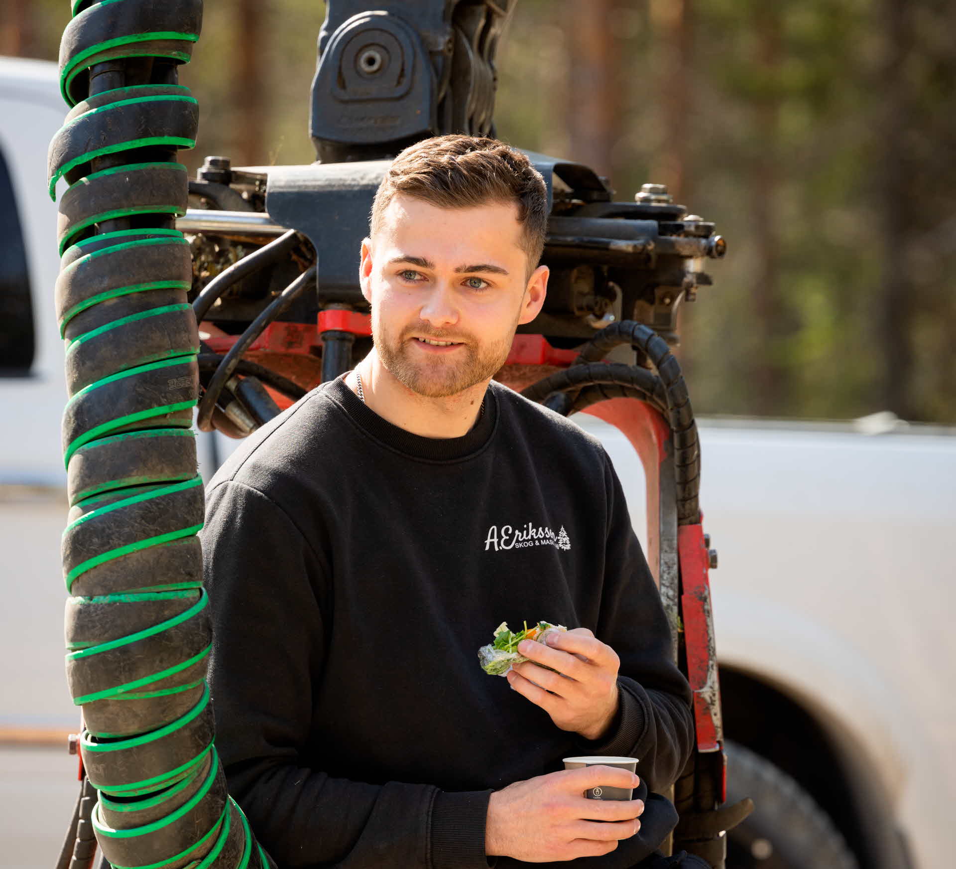 Albin äter en macka intill sin skogsmaskin.